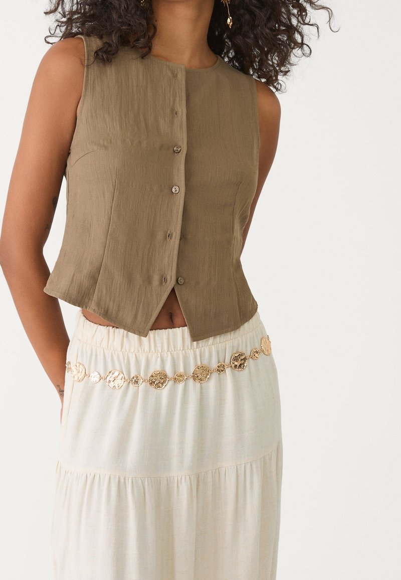 Femme portant un haut marron sans manches boutonné et une jupe crème avec une ceinture style pièce d’or autour de la taille.