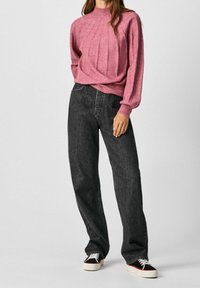 Růžový pletený svetr s ribovanou strukturou a vysokým límcem, doplněný volně střiženými tmavými džínami a černými teniskami s bílou podešví.