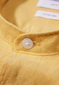 Camicia gialla con colletto realizzata in tessuto di cotone testurizzato, dotata di un bottone con quattro fori. Due etichette di cura visibili all'interno del colletto.