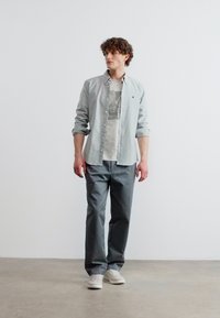 Νεαρός άνδρας όρθιος, φοράει ανοιχτό γκρι πουκάμισο πάνω από μπλούζα με στάμπα, σκούρα γκρι παντελόνια και λευκά αθλητικά παπούτσια, μπροστά σε απλό λευκό φόντο.