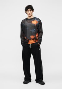 Jeune homme portant un t-shirt à manches longues en mesh noir avec un imprimé floral orange, un pantalon large noir avec un cordon blanc, et des chaussures noires.