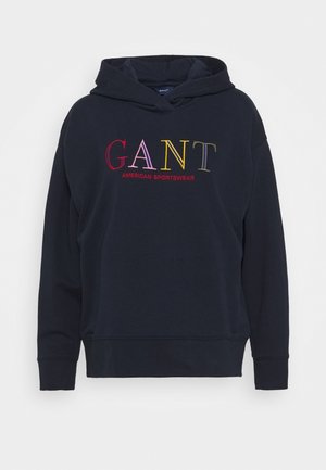 Laivansinis huppari, joka on valmistettu pehmeästä kankaasta ja jossa on edessä kengurupocket. Varustettu monivärisellä brodeeratulla tekstillä "GANT AMERICAN SPORTSWEAR."