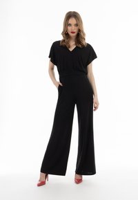 Schwarzer Jumpsuit mit V-Ausschnitt, weitem Bein und kurzen Ärmeln. Hergestellt aus einem weichen Stoff, mit tailliertem Schnitt und kombiniert mit roten High Heels.