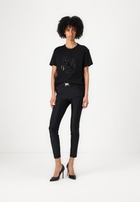 T-shirt nera con un design grafico in paillettes, abbinato a pantaloni neri a vita alta e tacchi a punta neri.