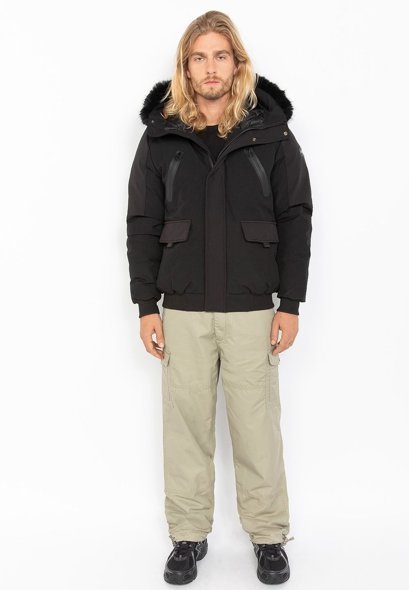 Schwarze isolierte Jacke mit Kunstfellkapuze, ausgestattet mit Reißverschlusstaschen, elastischen Bündchen und einer leicht taillierten Silhouette. Kombiniert mit hellen Cargo-Hosen.