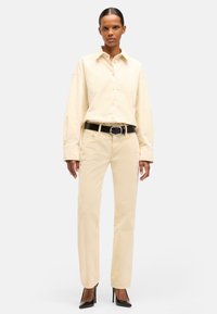 Camicia button-up color crema con maniche lunghe, abbinata a pantaloni beige chiaro a gamba dritta e una cintura nera, indossata con tacchi a punta neri.