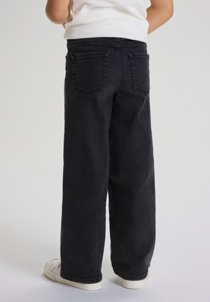 Barn stående iført sorte vidde bukser, hvide sneakers og en hvid skjorte, vist bagfra mod en ensartet lys baggrund.