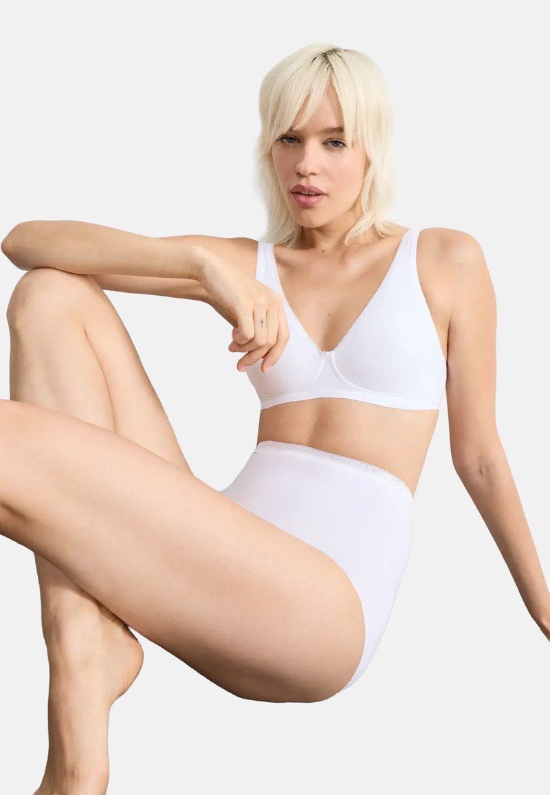 Femme aux cheveux blonds courts portant un soutien-gorge blanc et une culotte taille haute, assise avec une jambe pliée et le bras reposant sur le genou, sur fond blanc.
