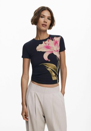 Navy blaues, geripptes kurzärmliges Hemd mit großem pinken Lilienmotiv und grünen Blättern auf der Vorderseite. Kombiniert mit hellen, gefalteten Hosen.