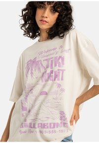 T-shirt oversize di colore crema con un design grafico viola che presenta palme, un tramonto e il testo "Benvenuti alla migliore TIKI NIGHT delle Hawaii."