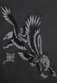 Due aquile ricamate dettagliate con le ali spiegate in nero e grigio su uno sfondo di tessuto scuro, accompagnate da un monogramma stilizzato.