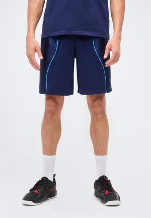 Uomo con pantaloncini sportivi blu navy con piping azzurro, scarpe da ginnastica nere e calzini bianchi a metà polpaccio, in piedi su uno sfondo bianco.