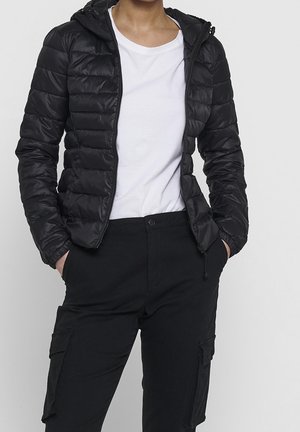 Person trägt eine schwarze gesteppte Kapuzenjacke über einem weißen T-Shirt und schwarze Cargohosen mit den Händen in den Taschen vor einem einfarbigen Hintergrund.