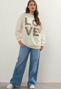 Felpa crema con la scritta "LOVE" in stampa leopardata, abbinata a jeans svasati blu e sneakers bianche. Vestibilità comoda, tessuto morbido, stile casual.