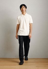 Λευκό πόλο πουκάμισο με γιακά, συνδυασμένο με μαύρο παντελόνι και μαύρα παπούτσια. Το μοντέλο περπατάει σε ξύλινο δάπεδο μπροστά από μια ουδέτερη πίσω μεριά.