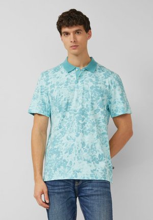 Młody mężczyzna ubrany w jasnoniebieską koszulkę polo w wzory i niebieskie dżinsy, stojący na tle jednolitego, jasnoszarego tła.