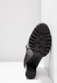 Botte noire à talon avec une semelle en caoutchouc texturée. Dispose d'une fermeture éclair latérale, d'une tige en cuir lisse et d'un talon cylindrique.