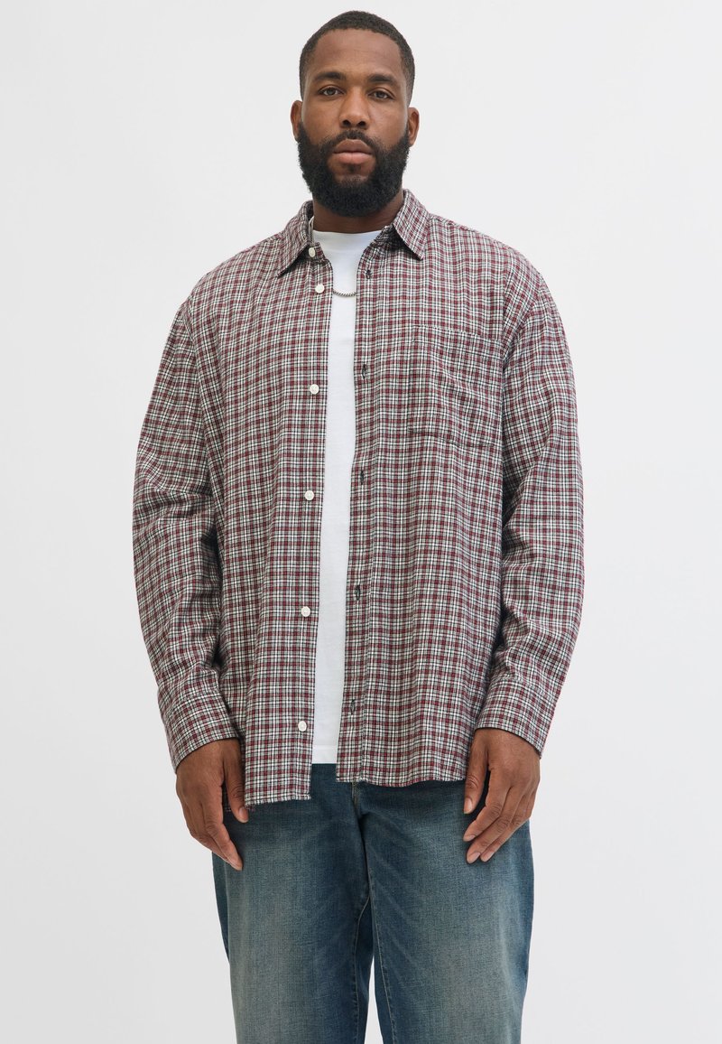 Homme avec une barbe portant une chemise à carreaux rouge et blanche boutonnée sur un t-shirt blanc et un jean bleu, sur un fond clair uni.