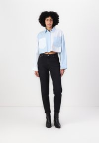Kraftigt beskuren skjorta med blå och vita ränder, matchad med högmidjade svarta jeans. Modellen bär svarta ankelboots och står mot en enfärgad bakgrund.