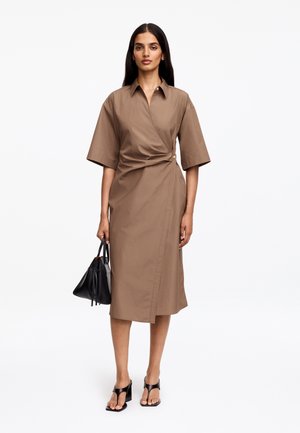 Femme portant une robe portefeuille marron mi-longue à manches courtes, tenant un sac à main noir et des sandales noires ouvertes à talons.