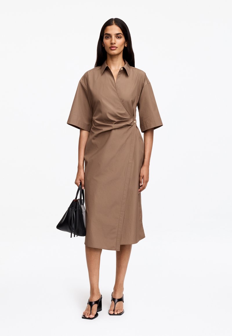 Femme portant une robe portefeuille marron mi-longue à manches courtes, tenant un sac à main noir et des sandales noires ouvertes à talons.