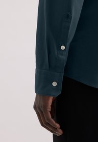 Maglietta a maniche lunghe color teal con bottoni bianchi, dotata di polsini con bottone e un design sagomato. Il materiale appare liscio con texture minima.