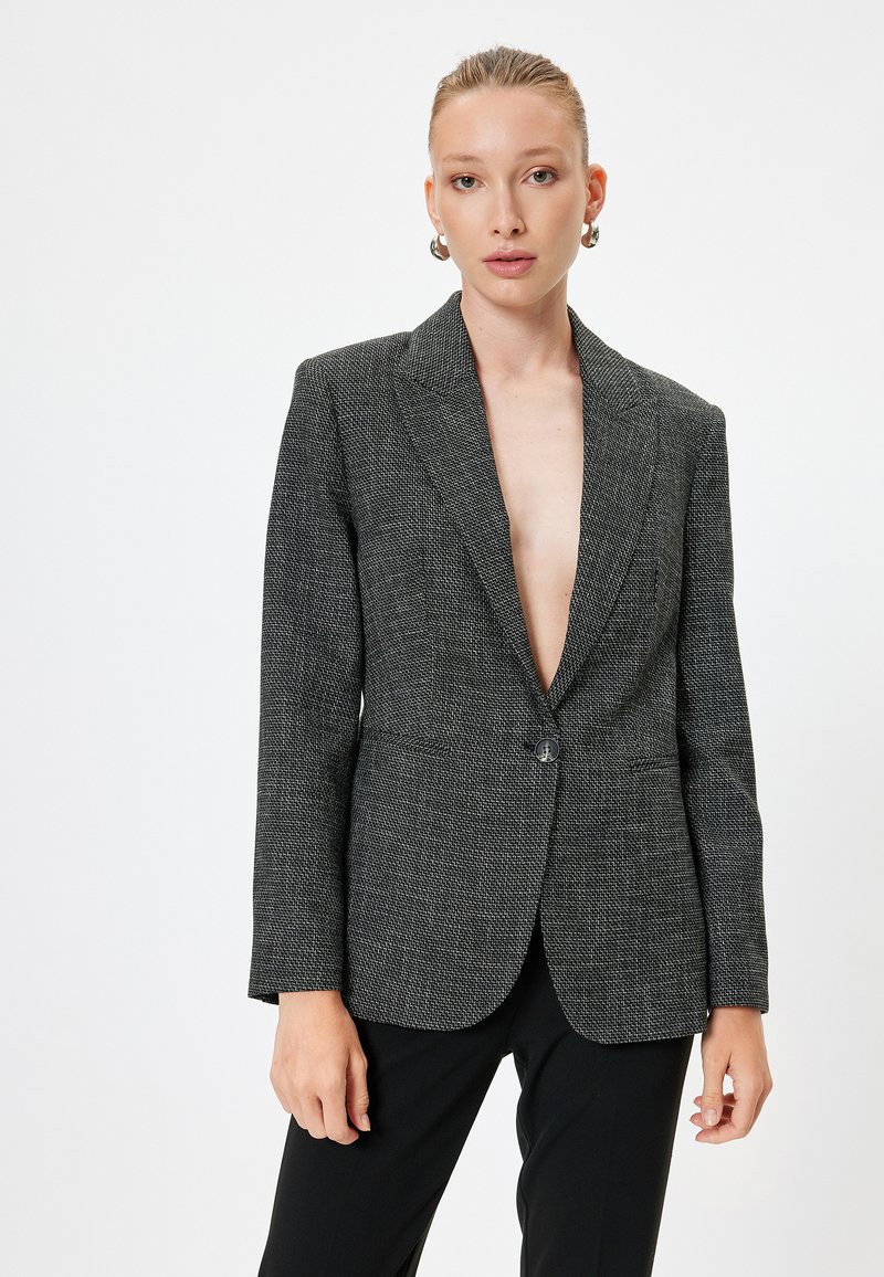 Blazer grigio con texture, a un bottone, con revers e due tasche frontali. Indossato sopra pantaloni neri dal taglio sartoriale.