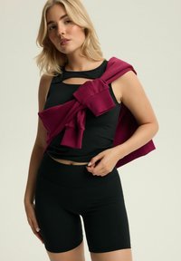 Czarny sportowy tank top z wycięciami w zestawie z czarnymi szortami. Bordowa bluza narzucona na jedno ramię. Gładka faktura, dopasowany krój.