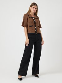 Mulher em pé contra um fundo branco, vestindo um cardigan de malha castanho com mangas curtas, com acabamentos e botões pretos, calças largas pretas e saltos altos pretos.