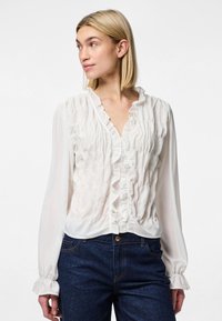 Blusa branca de mangas longas com decote franzido e detalhe na frente, apresentando pregas elásticas e tecido suave e translúcido, combinada com jeans de ganga escura.