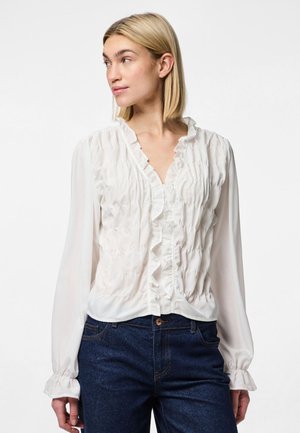 White, long-sleeve blouse with ruffled neckline and front, featuring elastic shirring and soft, sheer fabric, paired with dark denim jeans.