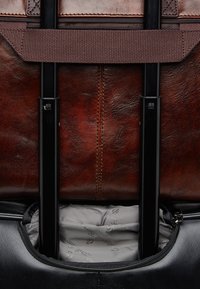 Brown leather bag with black accents, featuring sturdy black handles, a spacious interior, and a grey fabric lining with a subtle logo pattern.