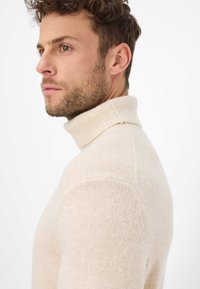 Maglione a collo alto beige realizzato in morbido materiale lavorato a maglia, con design aderente e colletto a coste, visto di profilo.
