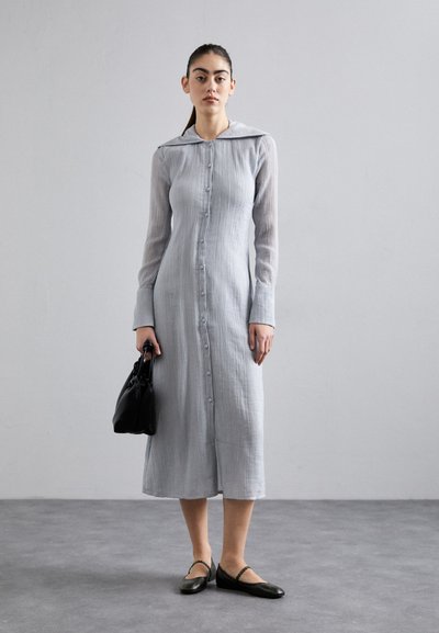 Vestido longo de manga longa em cinza claro, com colarinho e fecho frontal em botão, feito de um tecido texturizado; combinado com uma mala preta e sapatos planos escuros.