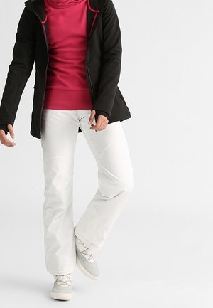 Persoon draagt een zwart jack, een rode hoodie, witte broek en grijze sneakers, staand tegen een effen lichte achtergrond.