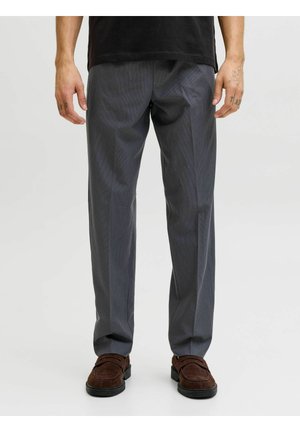 Homme portant un pantalon habillé gris et des mocassins en suède marron, debout avec les mains détendues le long du corps devant un fond uni.