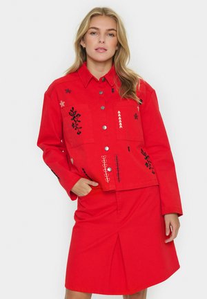 Giacca di jeans rossa con ricami floreali in bianco e nero, due tasche sul petto e bottoni a pressione argentati, abbinata a una gonna coordinata.