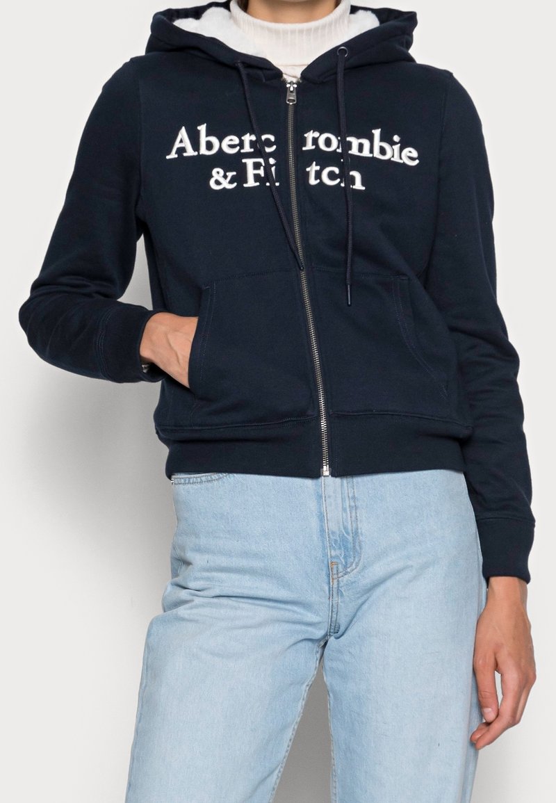 Felpa con zip colore navy con logo bianco "Abercrombie & Fitch". Presenta una tasca a marsupio frontale, cappuccio con cordino e fodera chiara a contrasto.