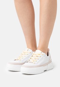 Baskets à plateforme blanches avec des accents rose clair, dotées d'une tige en cuir lisse, de semelles épaisses en caoutchouc et de lacets jaunes.