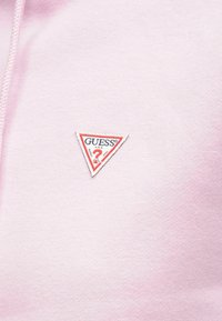 Ανοιχτό ροζ φούτερ με τριγωνική κόκκινη και λευκή ετικέτα λογότυπου «GUESS» στο στήθος. Μαλακό ύφασμα, casual σχέδιο, κουκούλα με κορδόνια.