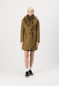 Oliven grøn uldfrakke med en faux fur krave, knaplukning og bælte i taljen. Båret over en sort kjole med sorte sneakers og hvide sokker.