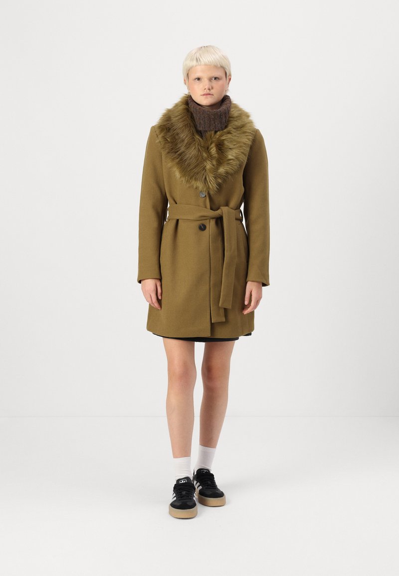 Oliven grøn uldfrakke med en faux fur krave, knaplukning og bælte i taljen. Båret over en sort kjole med sorte sneakers og hvide sokker.