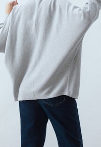 Grå, texturerad stickad tröja med avslappnad passform, med en tydlig söm längs ryggen, kombinerad med mörka jeans. Vit bakgrund.