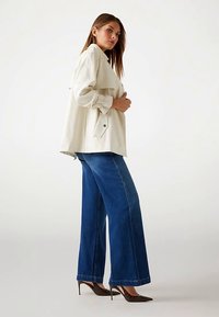 Cream-colored oversized jacket with flap pockets, paired with wide-leg blue denim jeans. Model wears brown textured heels.