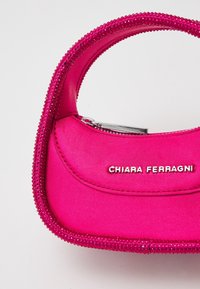 Różowa satynowa torebka o zaokrąglonym kształcie, ozdobiona akcentami przypominającymi klejnoty wzdłuż górnej krawędzi. Srebrny zamek błyskawiczny i detal z logo z przodu.