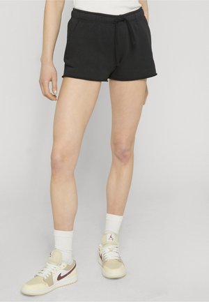 Persoon draagt zwarte shorts met koordje, witte sokken en beige sneakers met bordeauxrode logo's, staand tegen een lichte achtergrond.