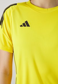 Geltonas sportinis marškinėliai trumpomis rankovėmis, apvaliu kaklu, su juodomis detalėmis pečių srityje ir juodu Adidas logotipu ant krūtinės. Lygi tekstūra.