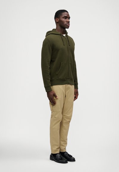 Homem vem com um casaco com fecho éclair verde oliva, calças de carga bege e mocassins pretos com franjas, de pé, virado para a direita, sobre um fundo simples.