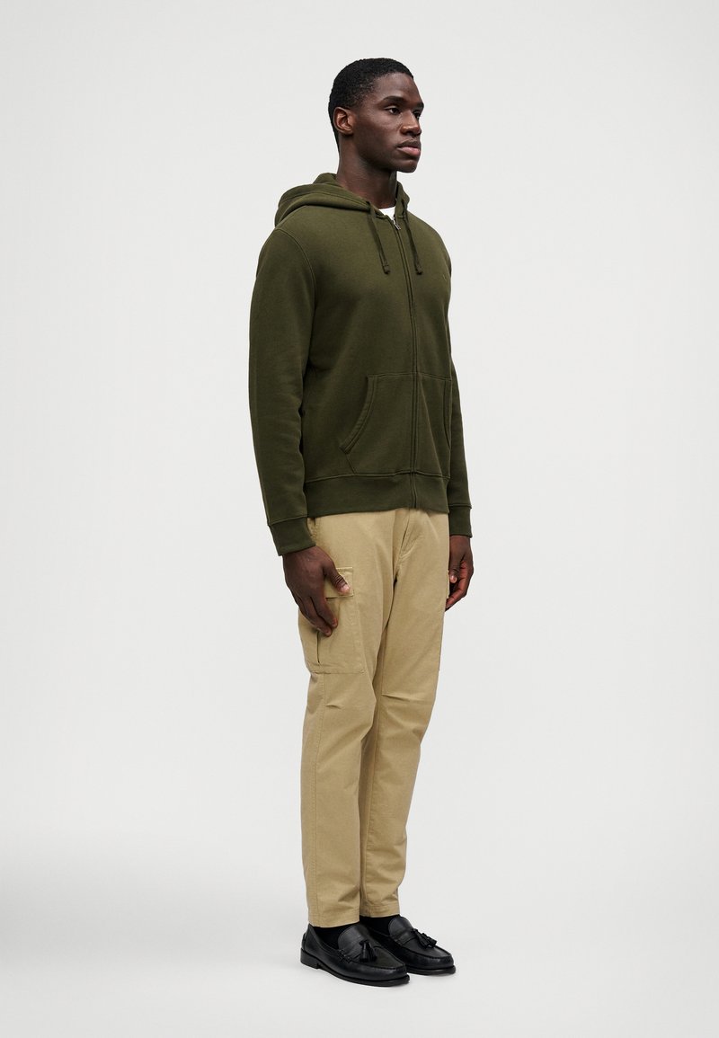 Hombre con una sudadera con cremallera de color verde oliva, pantalones cargo beige y mocasines negros con borlas, de pie mirando hacia la derecha sobre un fondo liso.