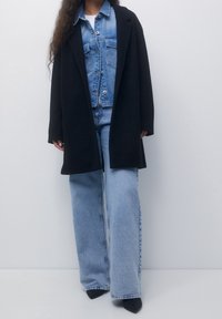 Mujer vistiendo un abrigo negro sobre una chaqueta de mezclilla y unos vaqueros de pierna ancha de color azul claro, con zapatos de tacón negro de punta afilada, contra un fondo liso.